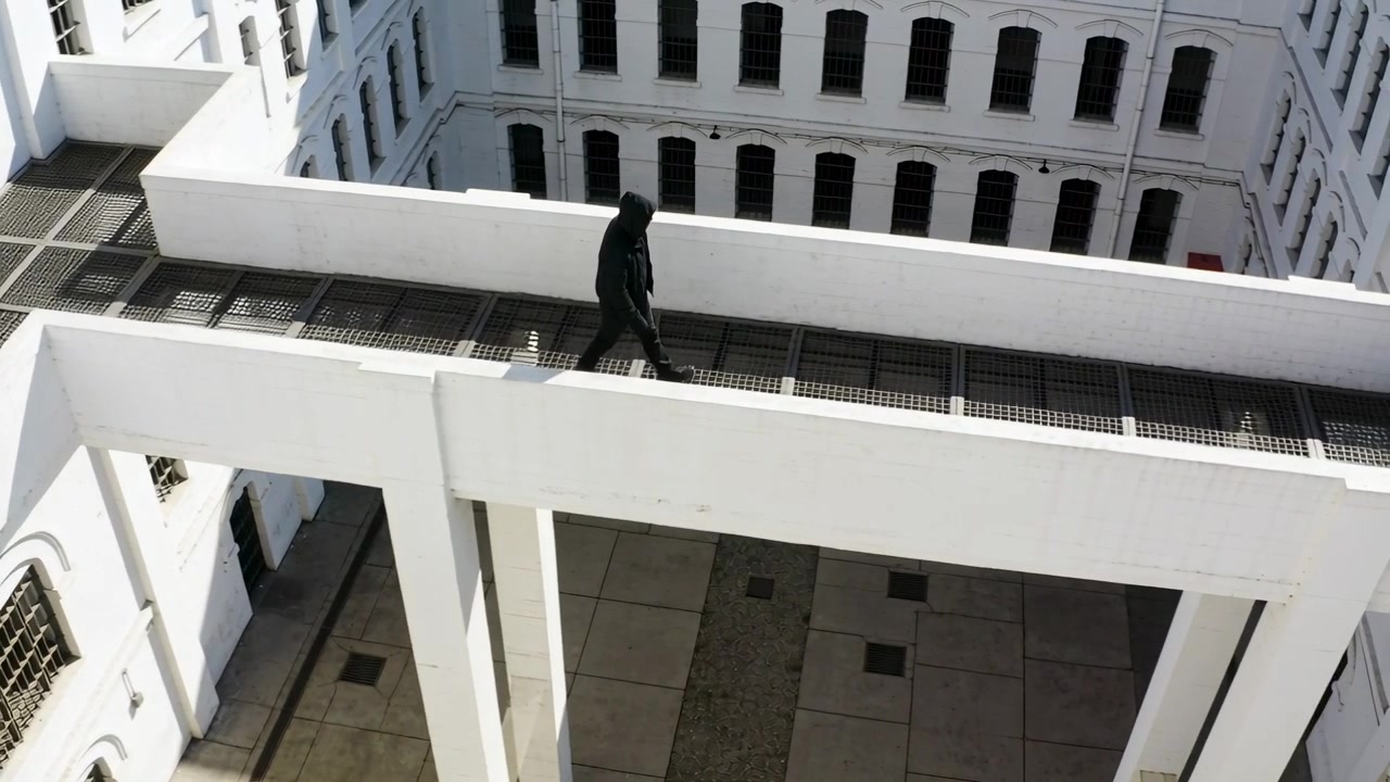 Mulai dari kiri, pria berjaket tudung berjalan di jalan setapak berjeruji di dalam halaman, melewati jendela berjeruji.