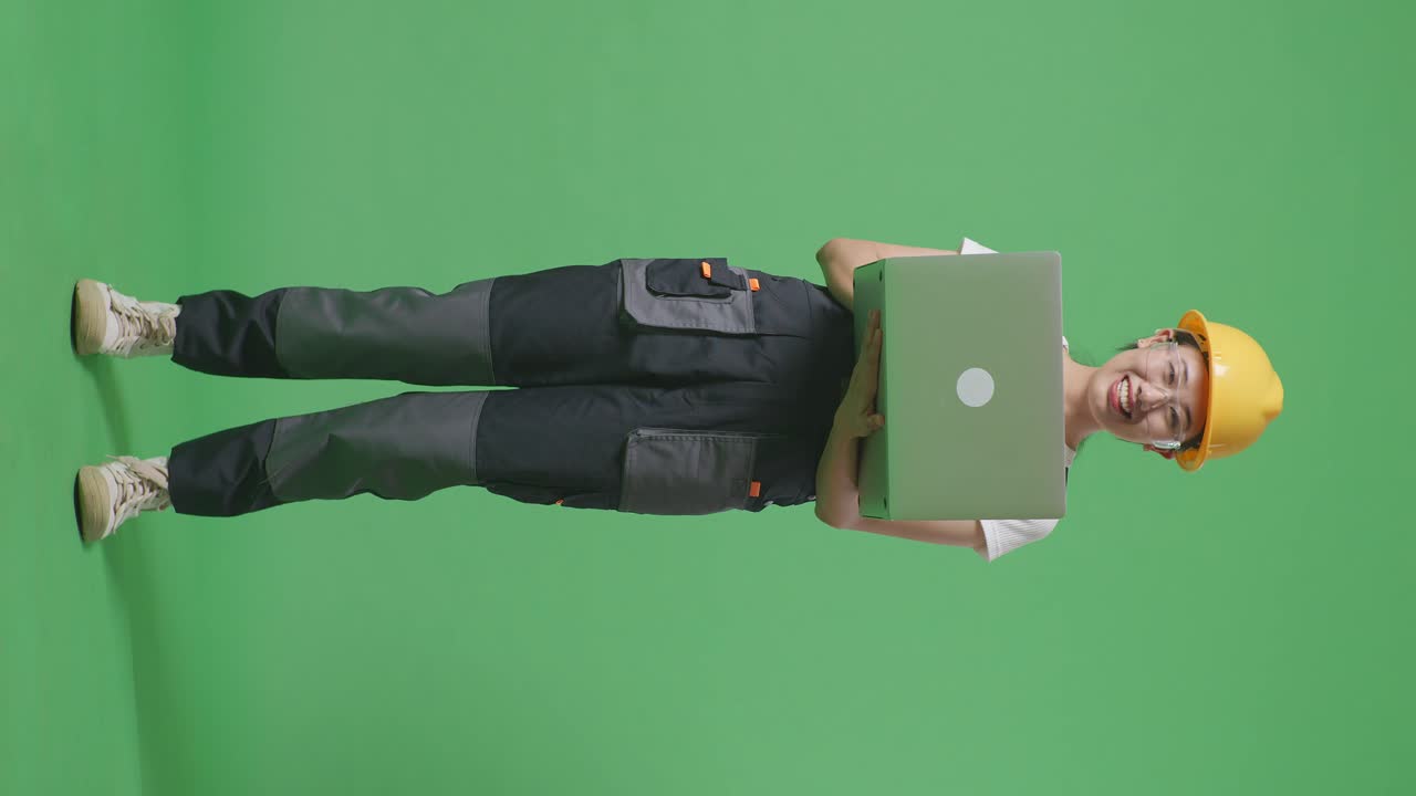 cuerpo lleno de trabajadora asiática que usa gafas de protección y casco de seguridad usando una computadora portátil y sonriendo mientras está de pie en el estudio de fondo de la pantalla verde