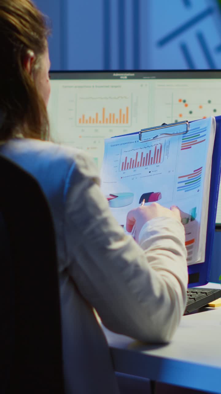 Vertical video: Back view of manager analysing graphics from clipboard at midnight