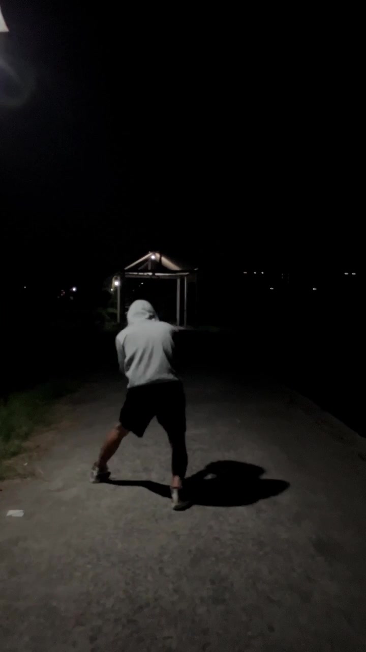 Vertical 4K tracking camera follow shot from behind a male athlete in hoodie walking and training along an empty road under street lights at dark night Cinematic fitness broll