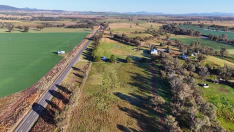 drone footage glides above a gravel road bordered by green fields, scattered trees, and rural homes under clear daylight in tamworth, australia