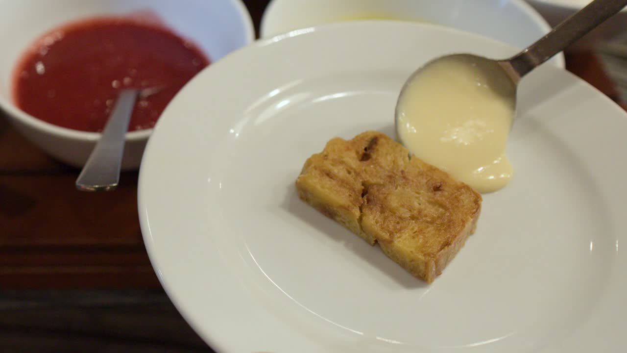 Una persona versa una cremosa salsa alla vaniglia da un cucchiaio su un budino di pane in un ristorante