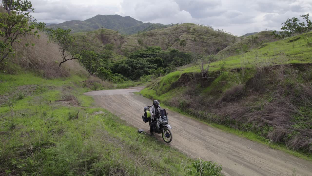 Drone shot a lone biker on a rugged dirt road, carving a path through a stunning mountainous landscape. Dramatic contrast of green and dry vegetation, deep connection with nature's raw beauty