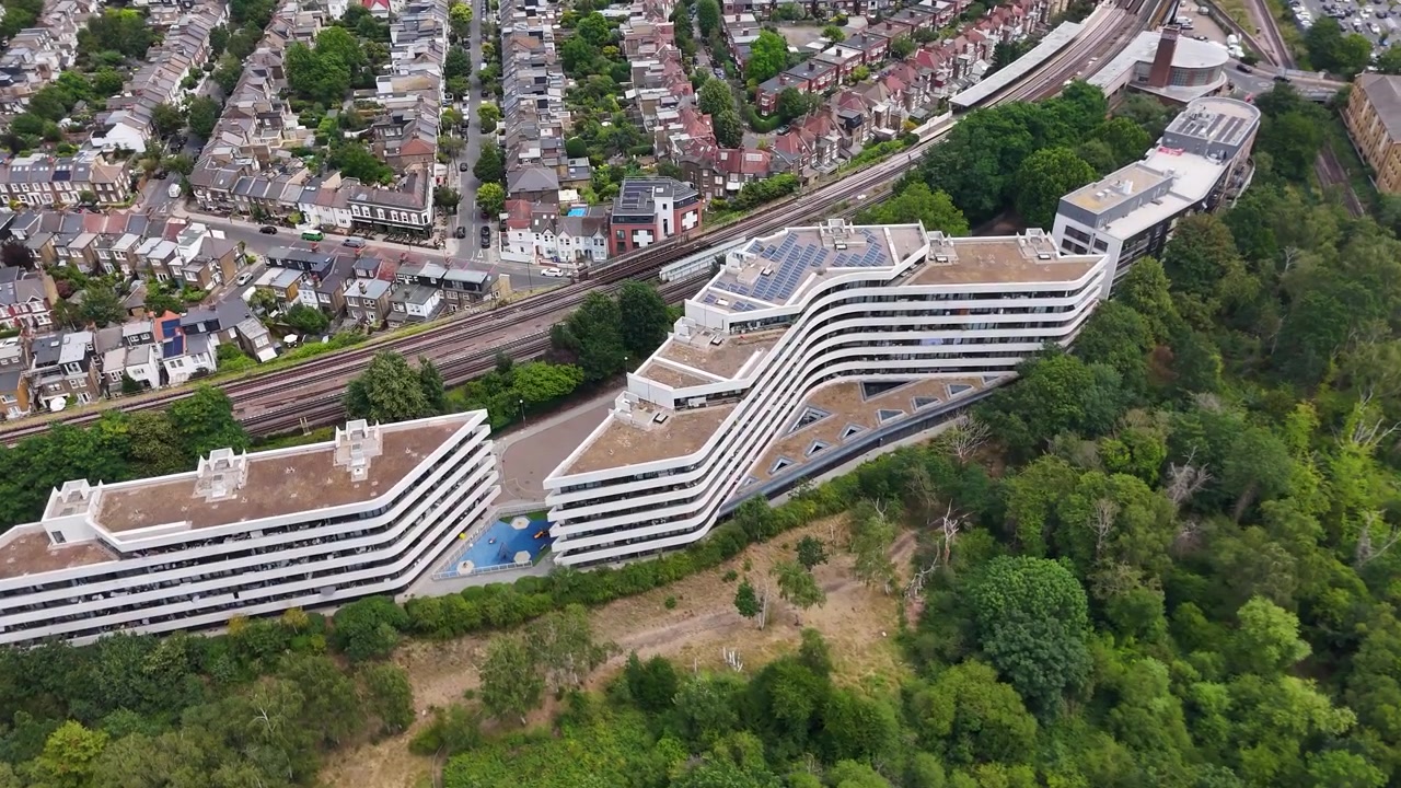Lungo edificio residenziale bianco e curvo chiamato Edmunds House che si snoda lungo il bordo di un taglio ferroviario di West London sopra alberi maturi