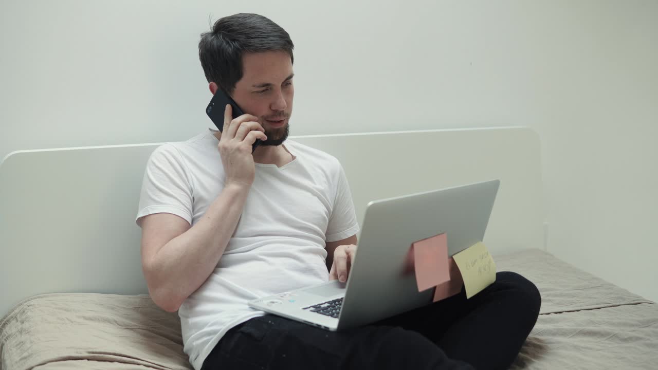젊은 남자가 스마트폰으로 이야기하고 아파트에서 노트북 모니터를 보고 있습니다.