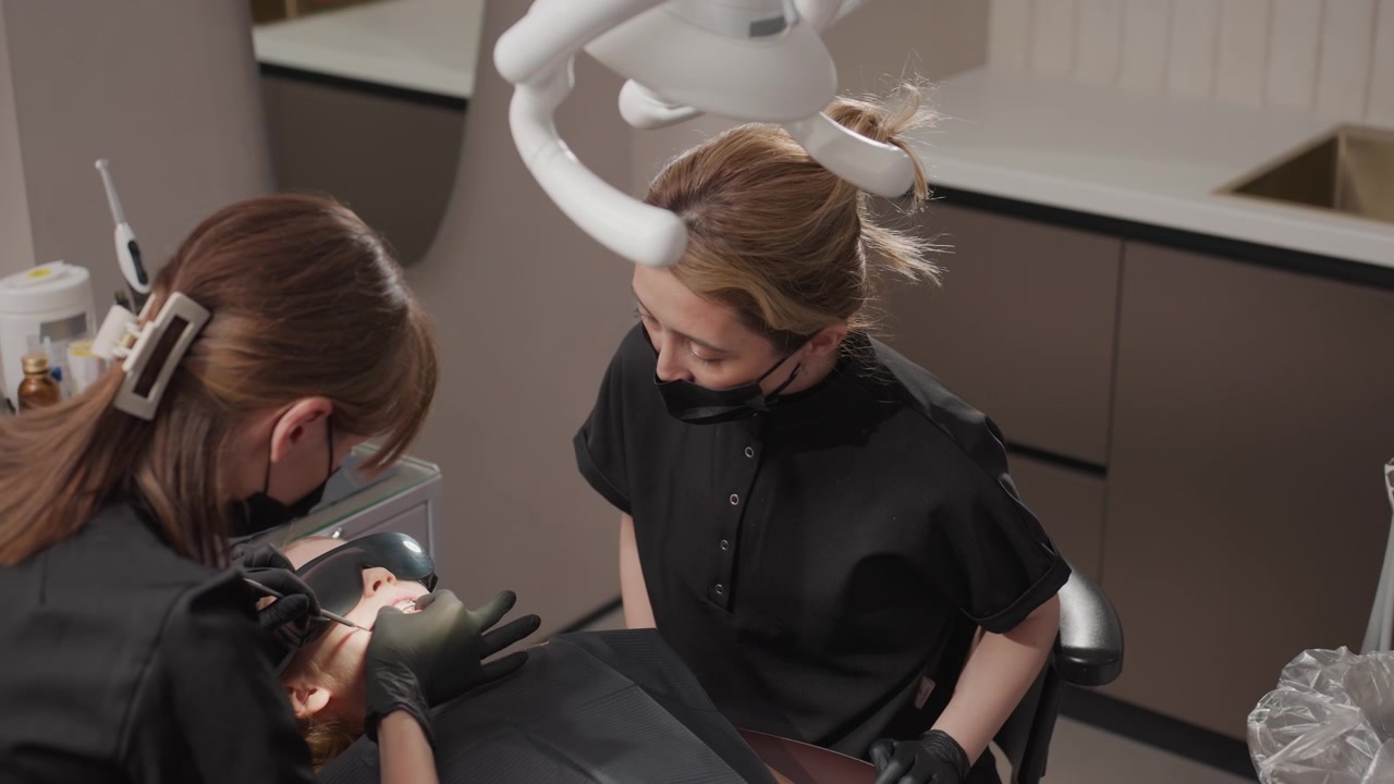 Primo piano di due professionisti dentali che eseguono una procedura ortodontica su un paziente in una clinica moderna. Strumenti e attrezzature dentali visibili, inclusa un'area di esame illuminata e attrezzature mediche