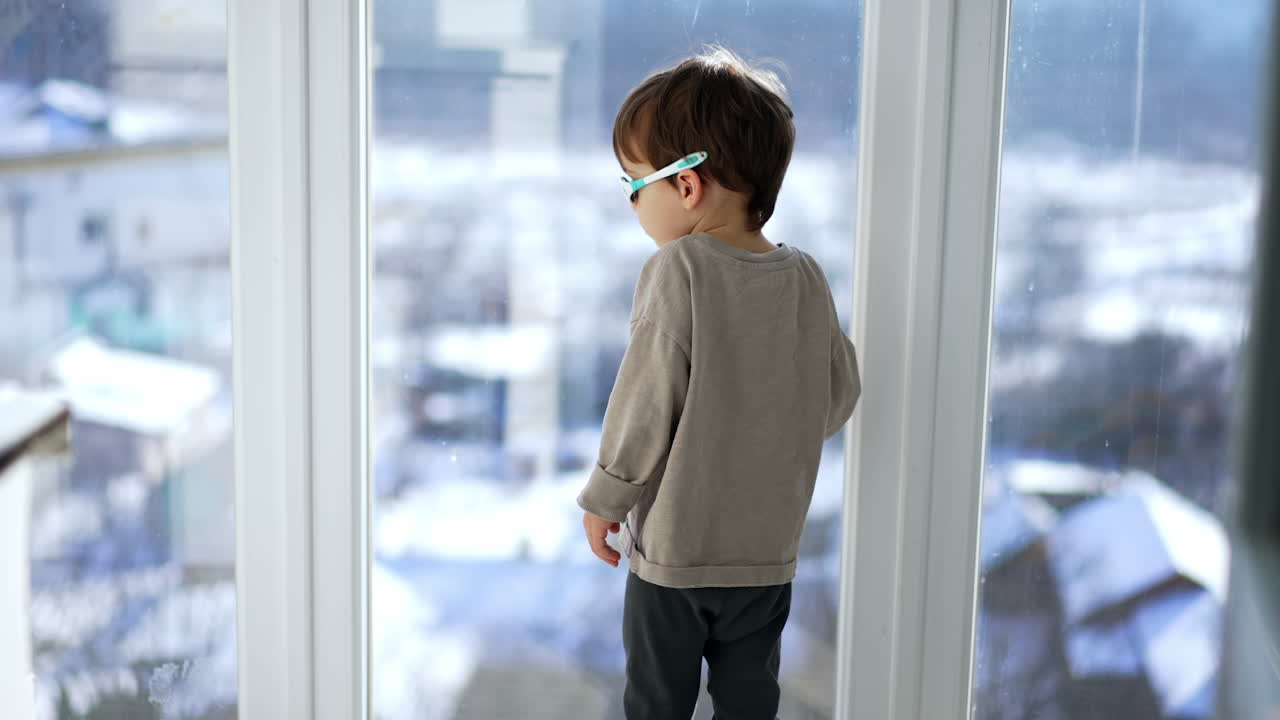 Caucasian baby boy wearing grey sweater and sunglasses comes up to a window. Kid turns to camera and looks at black cat jumping up.