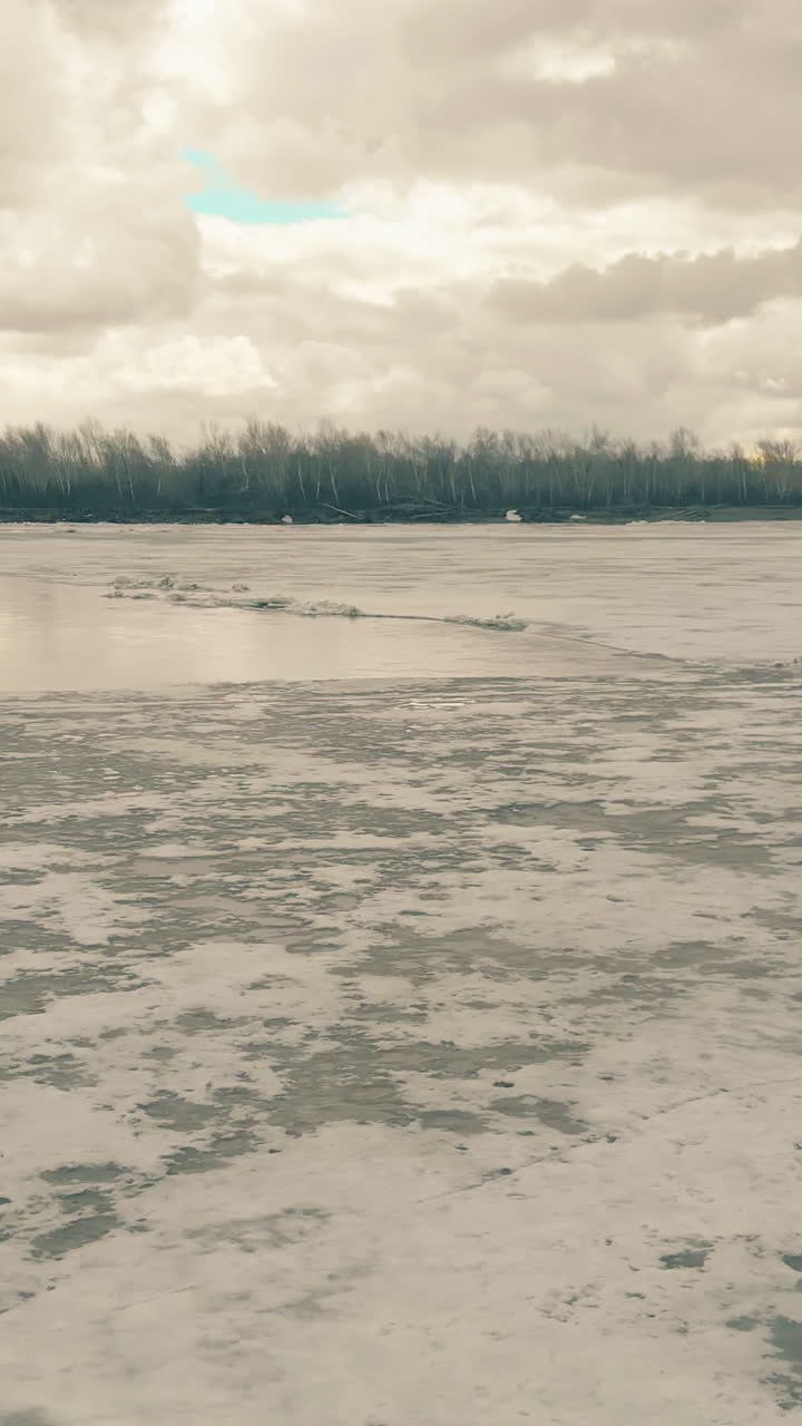 pictorial frozen river with patch of open water in ice against bank with dense forest bridge under cloudy sky upper view