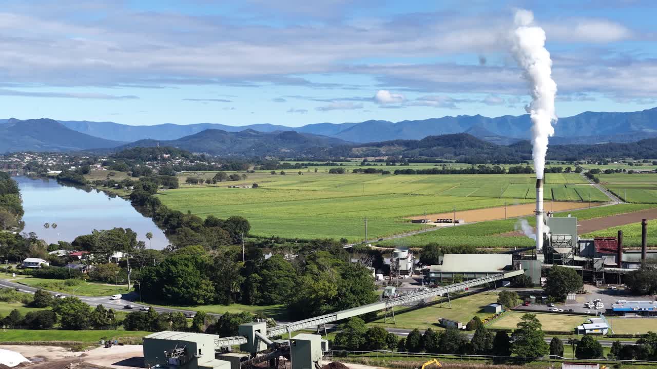 Aerial view of a sugar mill with lush green fields and distant mountains under clear skies