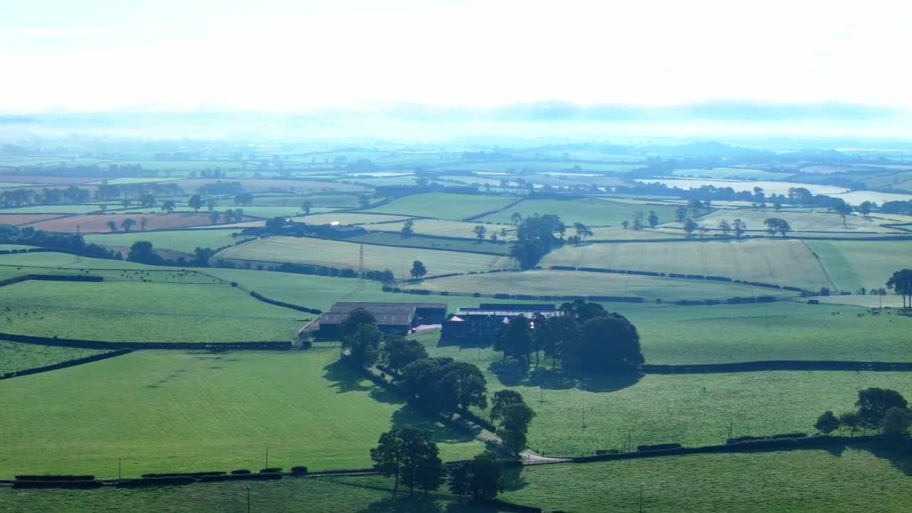 Aerial view of misty farmland and rolling countryside at dawn. Light fog hangs over fields, hedgerows, and country roads in a tranquil rural morning landscape