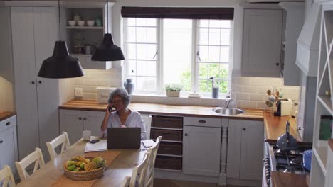 Mujer-Afroamericana-Mayor-Usando-Una-Computadora-Portátil-Y-Hablando-Por-Teléfono-Inteligente-En-Casa