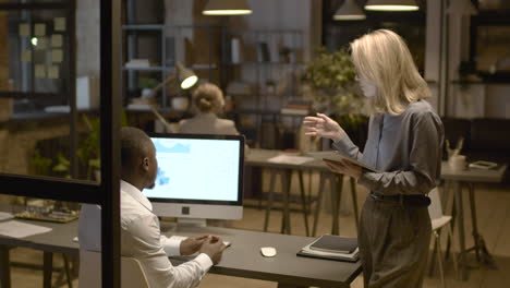 rear view of american man sitting at desk while talking with female coworker who is holding a tablet in the office 1