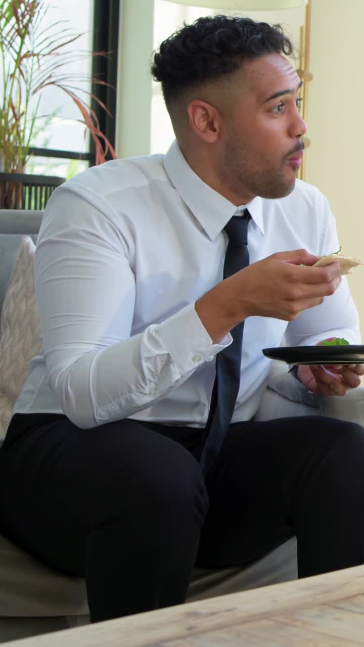 Vertical video: Leaning, man in tie adjusting small black plate, snacking on avocado toast on sofa