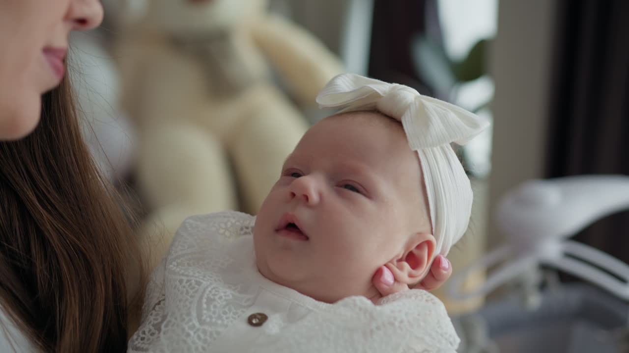 Primo piano di una madre che tiene delicatamente in braccio una neonata vestita con un abito di pizzo bianco e una fascia per capelli, promuovendo