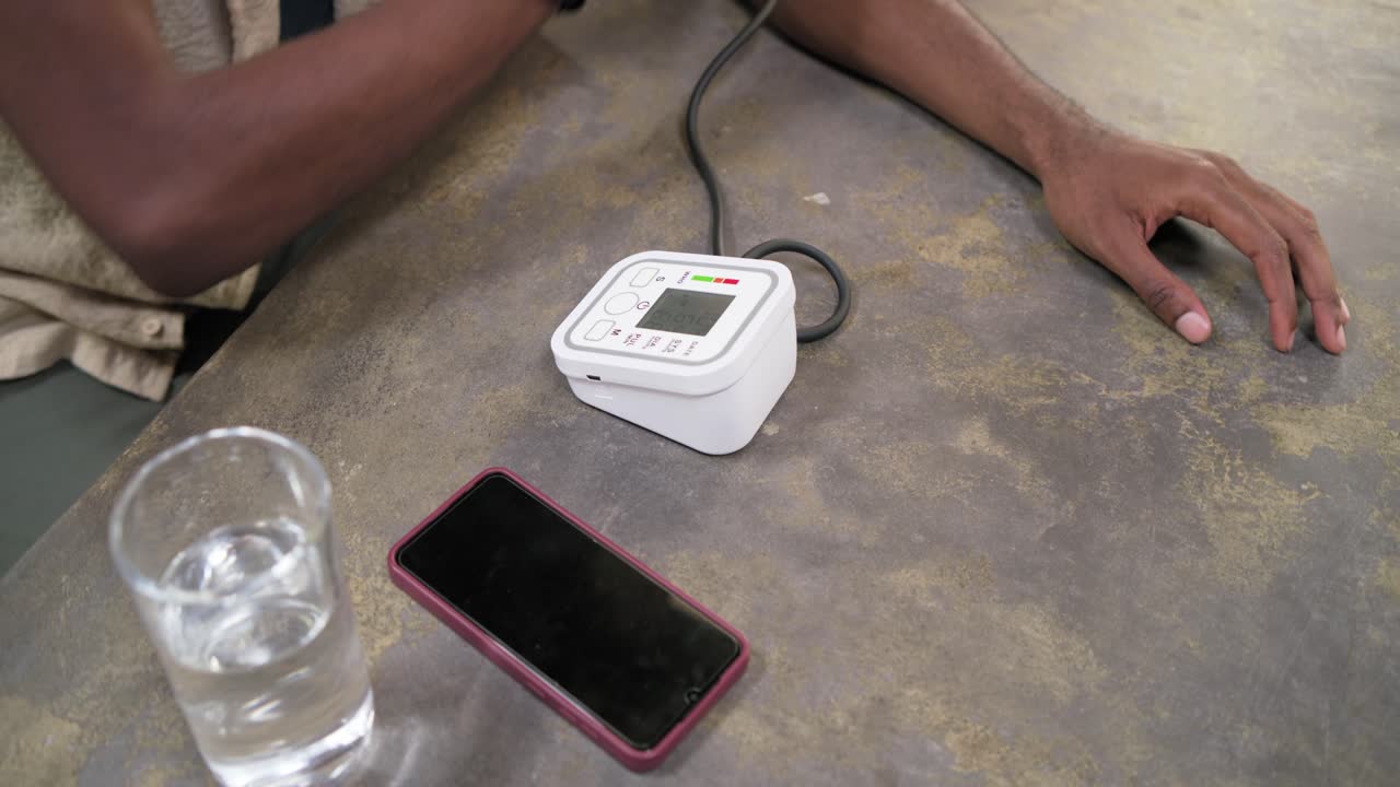 Uomo adulto africano che osserva il monitor, si rimbocca la manica, applica il bracciale e controlla la pressione sanguigna al tavolo.