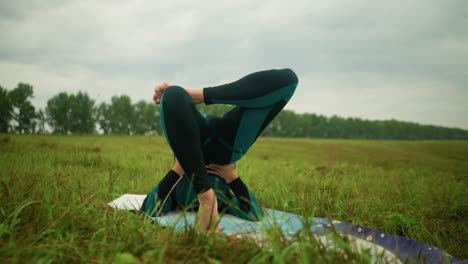 vista trasera de una mujer blanca acostada en una alfombra de yoga practicando la postura del puente con las manos apoyando su cintura en un campo cubierto de hierba bajo un cielo nublado, los árboles están alineados en la distancia
