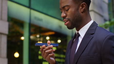 Young-Businessman-Wearing-Suit-Talking-On-Mobile-Phone-Using-Built-In-Microphone-Standing-Outside-Offices-In-The-Financial-District-Of-The-City-Of-London-UK-Shot-In-Real-Time-1