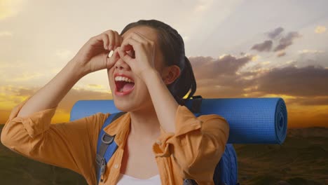 close up of asian female hiker with mountaineering backpack making binoculars gesture looking at something then saying wow while standing on the top of mountain during sunset time