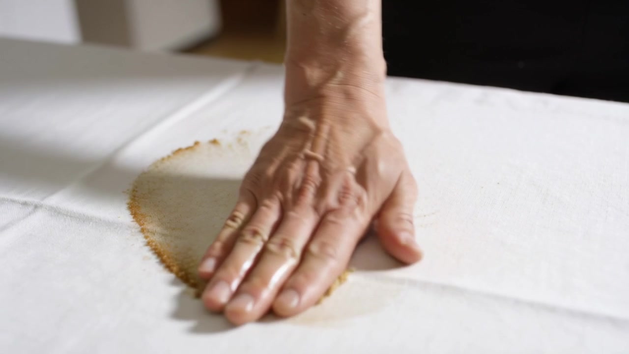 Mão que entra pressionando, esfregando mancha marrom em pano branco na mesa, enxugando o derramamento, manga escura.