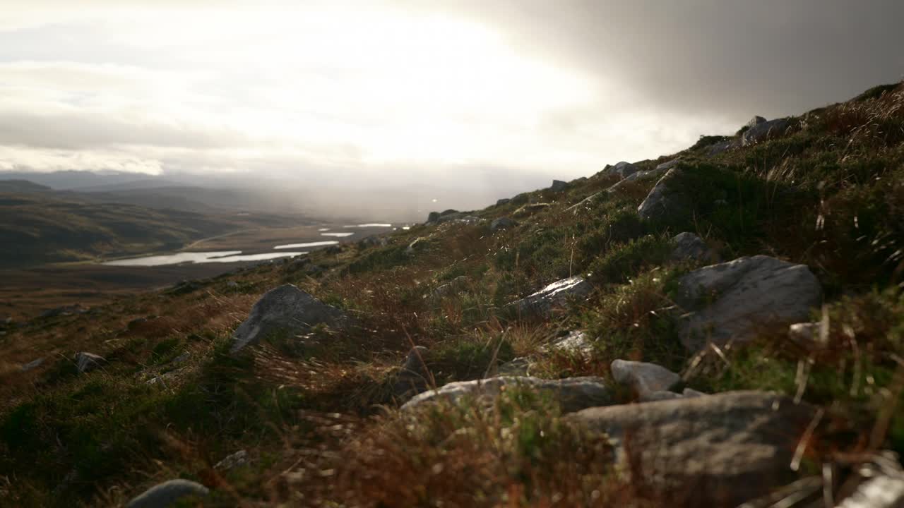 los vientos rápidos soplan hierba en primer plano contra un paisaje de montañas y lagos en las tierras altas del noroeste de escocia mientras rayos de luz perforan el cielo