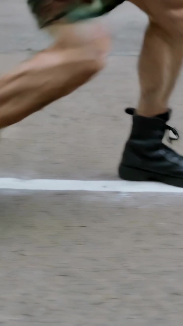 Video verticale: Sollevando il piede destro, un corridore corre, mantenendo il passo lungo la linea della corsia, con pantaloncini mimetici e stivali.