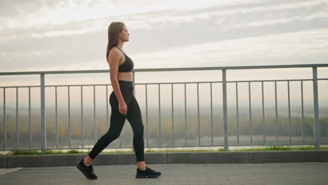 Pretty woman in black leggings walking majestically beside iron fence, looking out into the distance, the background features a serene outdoor scene with clear skies and distant views