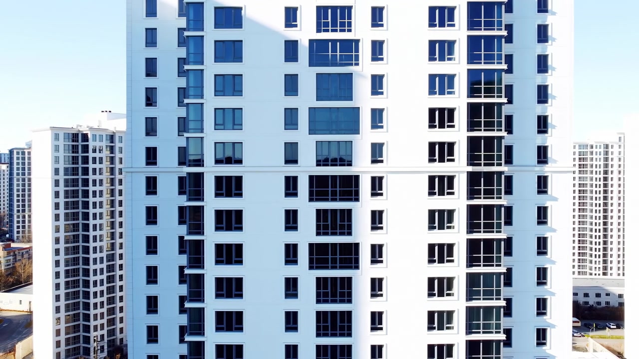 La cámara se mueve lateralmente pasando por un edificio residencial iluminado por el sol con fachada blanca, ventanas oscuras y balcones apilados, mientras torres de apartamentos distantes permanecen visibles contra un cielo azul despejado