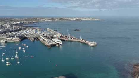 St-Peter-Port-Guernsey-flight-over-ferry-terminal-with-ferry-in-dock,-commercial-dock-with-cranes-with-QE-II-Marina-in-background-and-views-over-Belle-Greve-Bay-on-bright-sunny-day