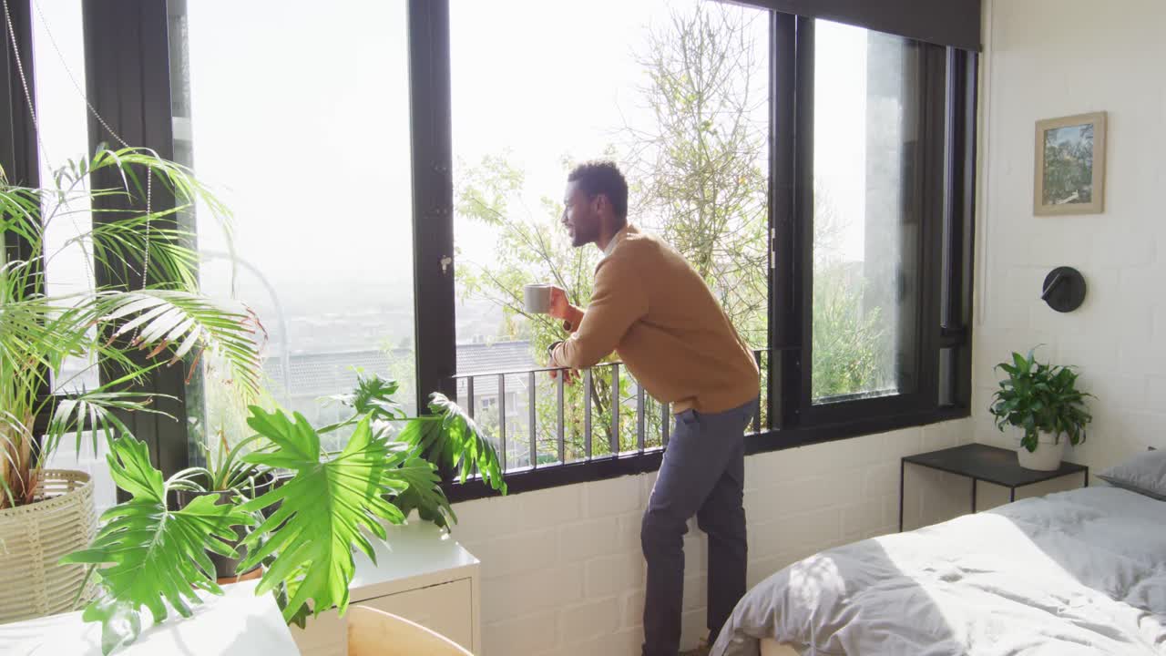 feliz hombre afroamericano mirando a través de la ventana y bebiendo café