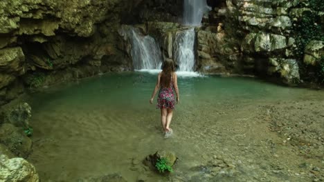 scenic landscape view above young caucasian woman sitting on rock in shallow water, stands up and walks towards clear fresh yahshoush mountain waterfall, lebanon, behind overhead drone pull back