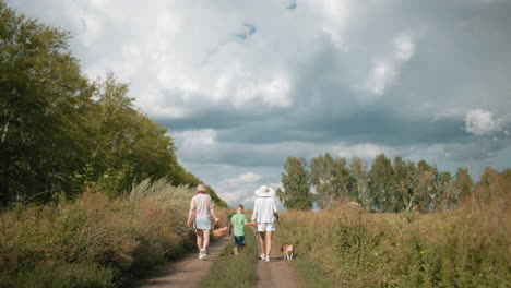 rear view of family walking on dirt road surrounded by lush greenery during retreat, carrying picnic basket while child holds umbrella and dog on leash under partly cloudy sky