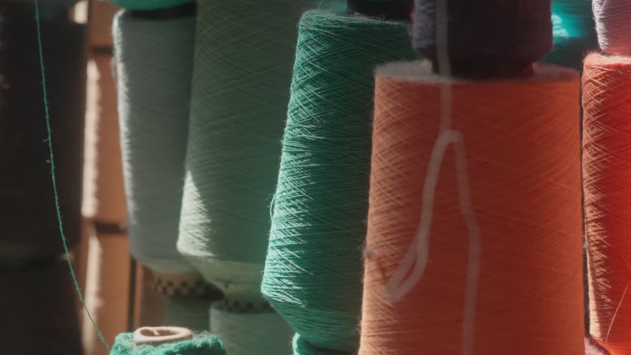 Zoomed-out view of colorful yarn cones in shades of green, orange, turquoise, and more, lined up with loose threads in a traditional textile weaving mill