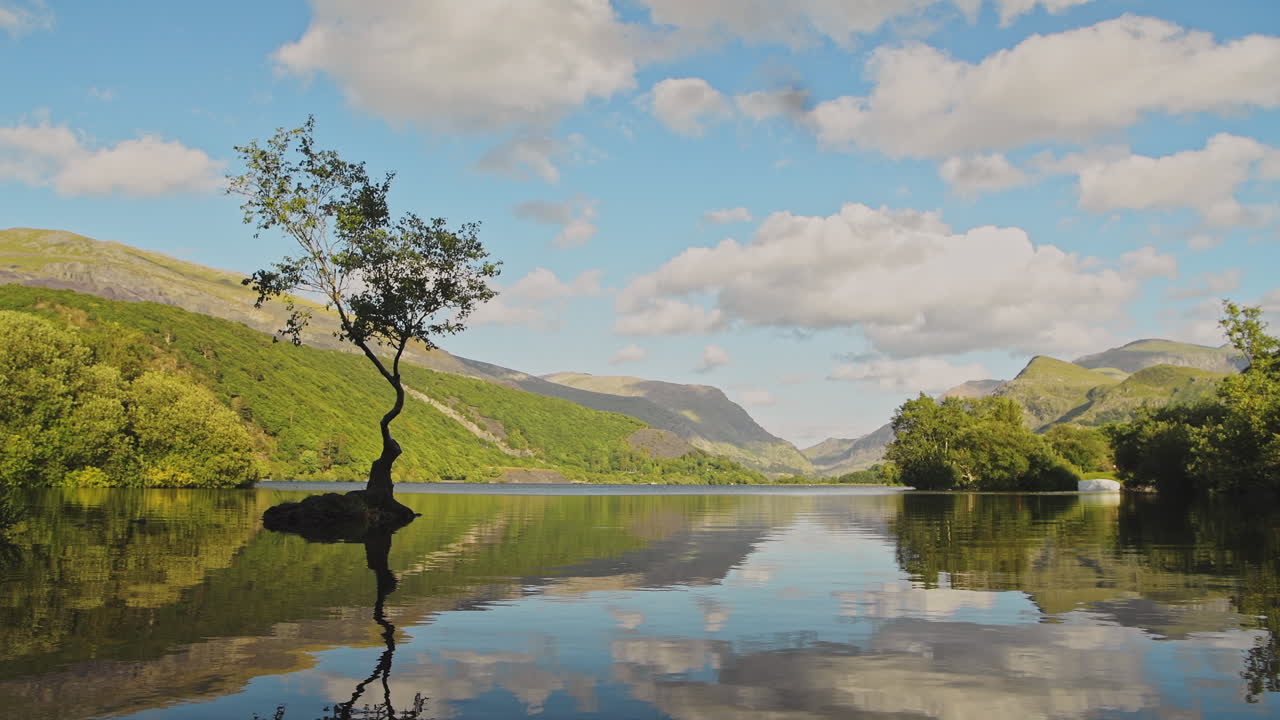 Clouds reflecting on lakes surface with beautiful british landscape reflections of lone tree on beautiful sunny day at Llyn Padarn Lake, Wales