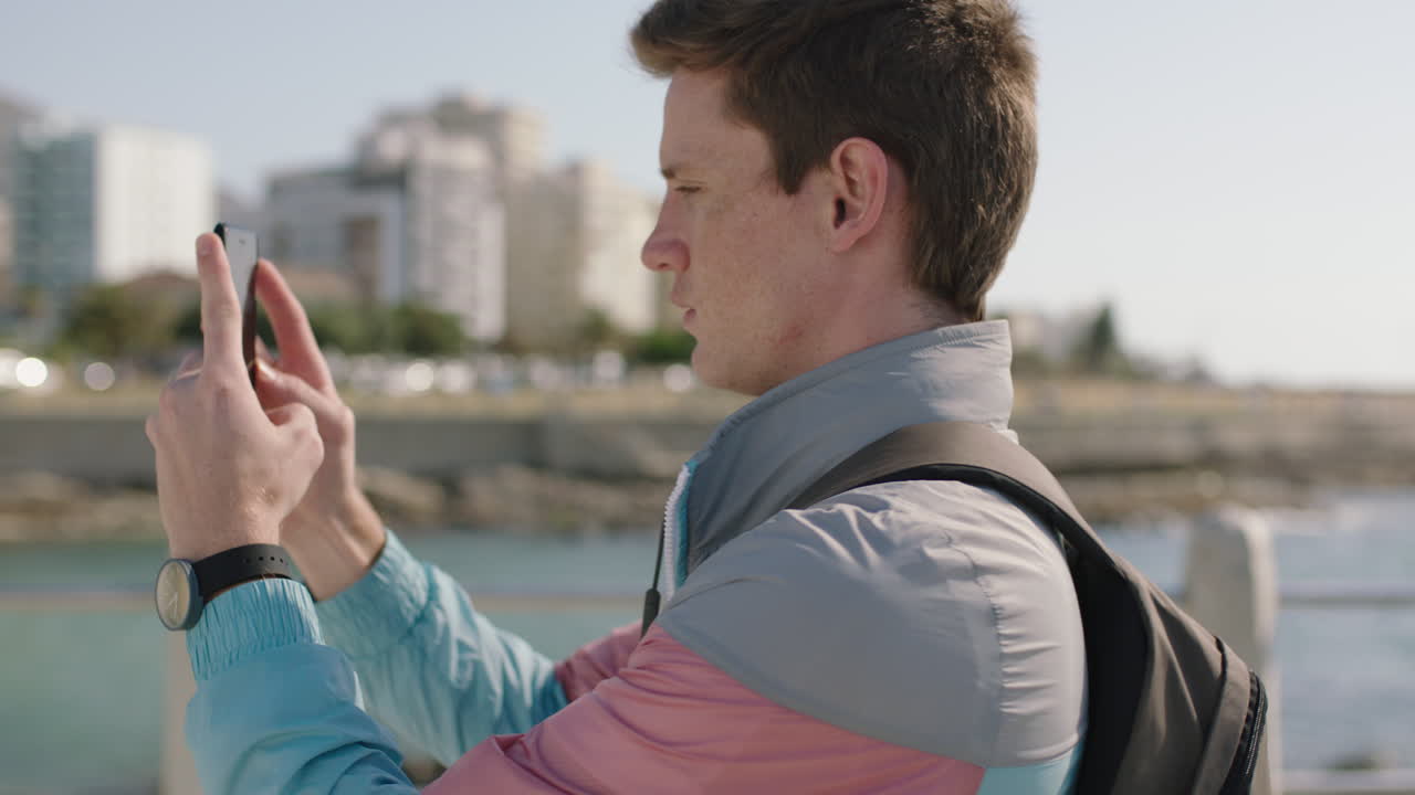 retrato de un atractivo hombre joven de cabeza roja usando un teléfono inteligente tomando fotos en una playa soleada