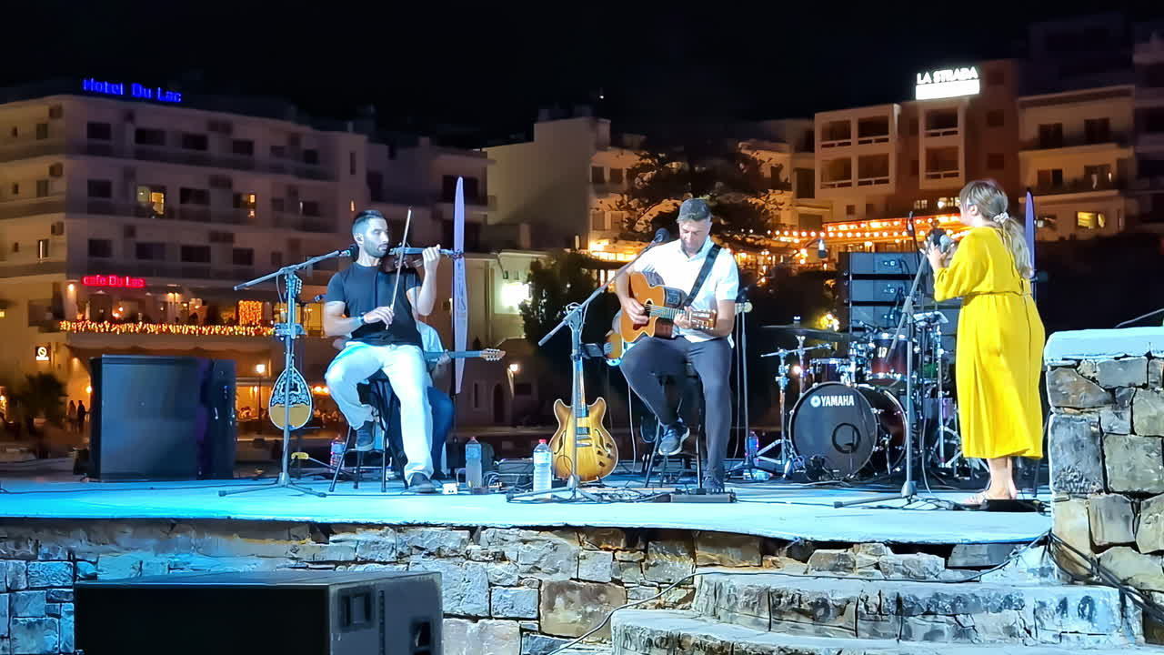 A live folk band performs on an outdoor stage at night in Agios Nikolaos under city lights
