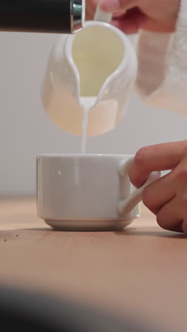 una mujer con túnica de terry agrega espuma lechosa a una taza de porcelana de una jarra disfrutando temprano en la mañana.
