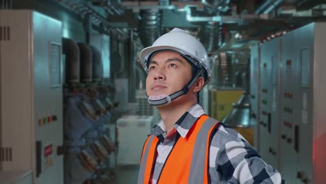 close up side view of asian male engineer wearing safety helmet looking around while standing with arms akimbo in engine control room, work of electrical generators