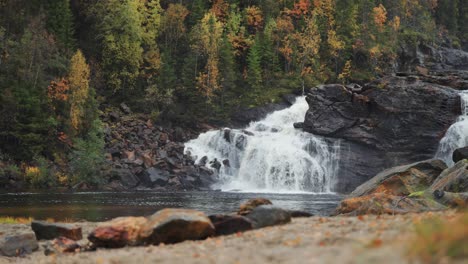 A-majestic-waterfall-cascades-over-dark-rocks,-overlooking-a-serene-lake-and-sandy-beach-below,-creating-a-captivating-scene-of-natural-beauty