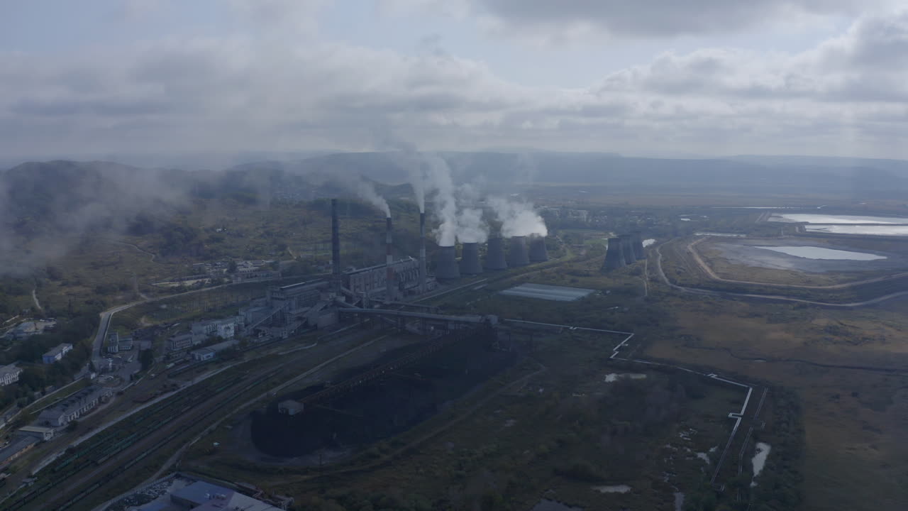 Lenta carrellata all'indietro di una centrale elettrica a carbone con i suoi camini e ciminiere che rilasciano fumo bianco e grigio nell'aria in una giornata soleggiata e nuvolosa.