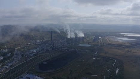 slow push back shot of a coal-fired power station with its chimneys and funnels releasing white and grey smoke into the air on a sunny and cloudy day