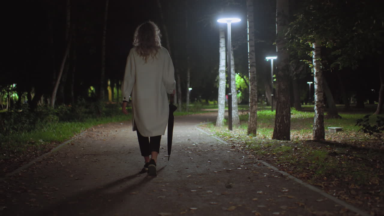Back view of young lady in long white coat holding umbrella walking alone on quiet park path illuminated by streetlights, surrounded by trees and fallen leaves in calm nighttime atmosphere
