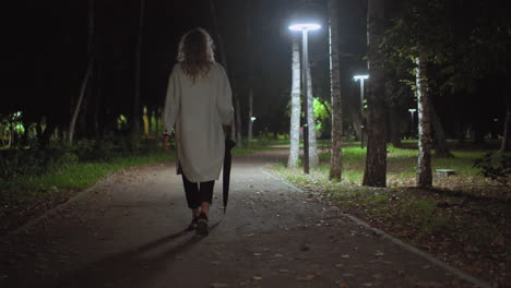 back view of young lady in long white coat holding umbrella walking alone on quiet park path illuminated by streetlights, surrounded by trees and fallen leaves in calm nighttime atmosphere