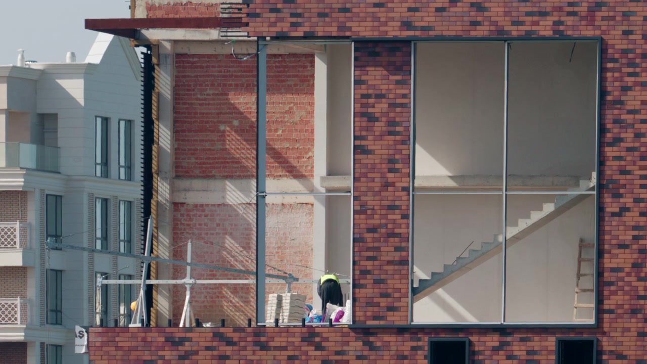 Lavoratori all'interno di un moderno edificio in costruzione, con pareti in mattoni a vista, interni grezzi e ampie aperture finestrate.