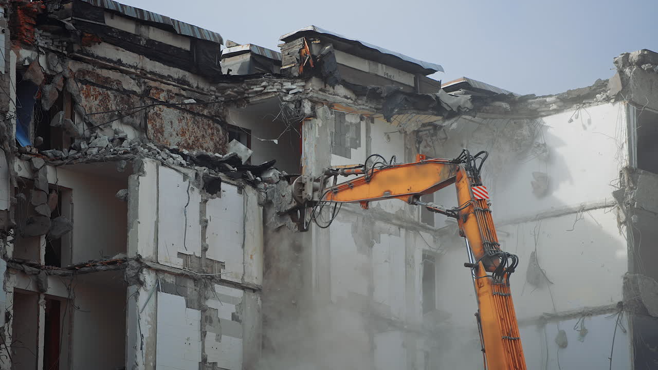 Hydraulic crasher of an excavator machine demolishing the house. High building wrecked by an earthquake being destroyed by a special machinery. Dust spreads all over the place.