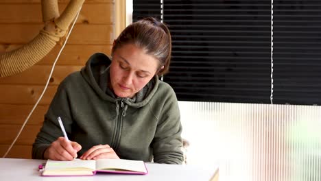 Young-woman-writing-in-journal-sitting-at-table-on-porch,-medium-shot