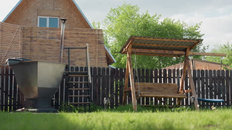 peaceful backyard scene showing wooden swing under canopy, metal outdoor hot tub with chimney and steps, dark fence, wooden privacy screen, lush green grass
