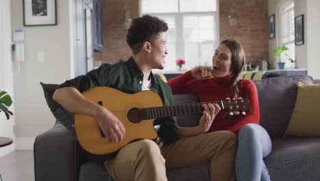 happy biracial couple sitting on sofa in living room playing guitar