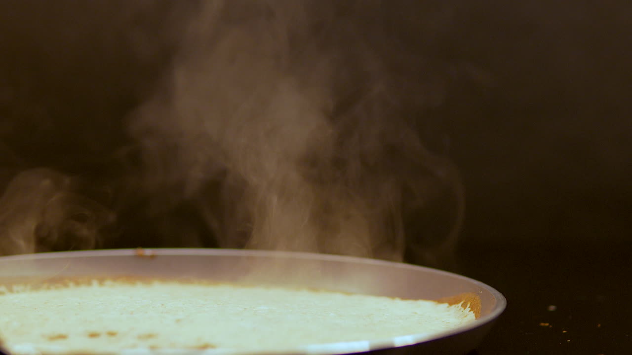 Close up panekuk tipis atau crepe yang sedang dimasak di atas wajan dengan uap putih tebal mengepul dengan latar belakang gelap. Sempurna untuk konsep kuliner dan sarapan