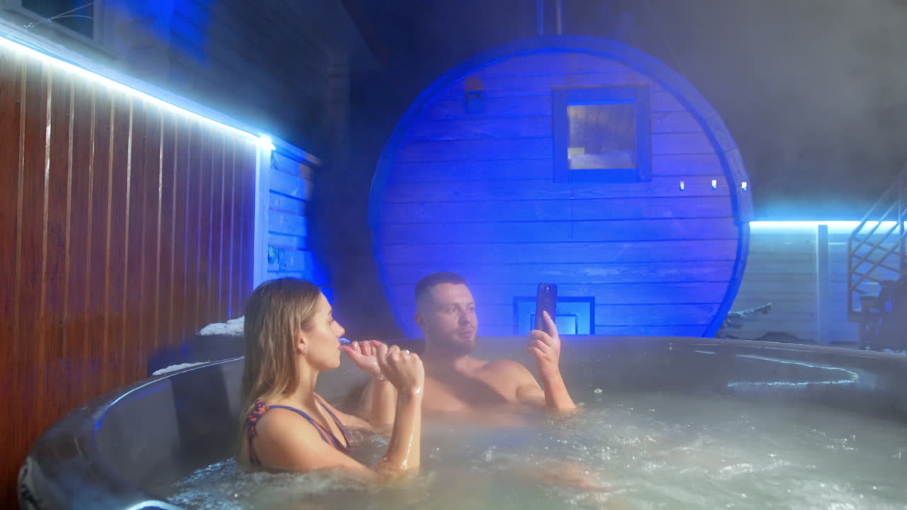 Caucasian couple sit in large hot tub in the yard. Woman is fixing her hair and man looks at his phone.