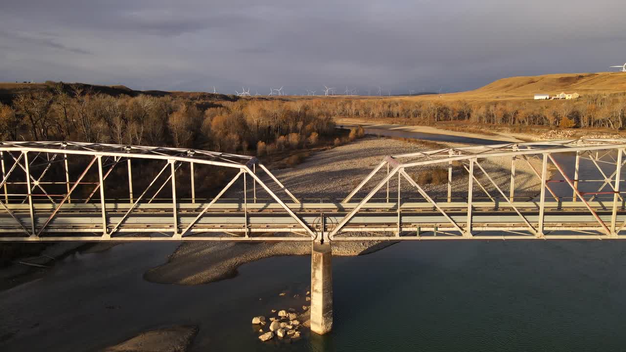 Drone approaching and flying over an old steel bridge during sunrise gloom in southern Alberta. Sustainable wind turbines underneath a dramatic cloudy sky in the background. 4k aerial footage.